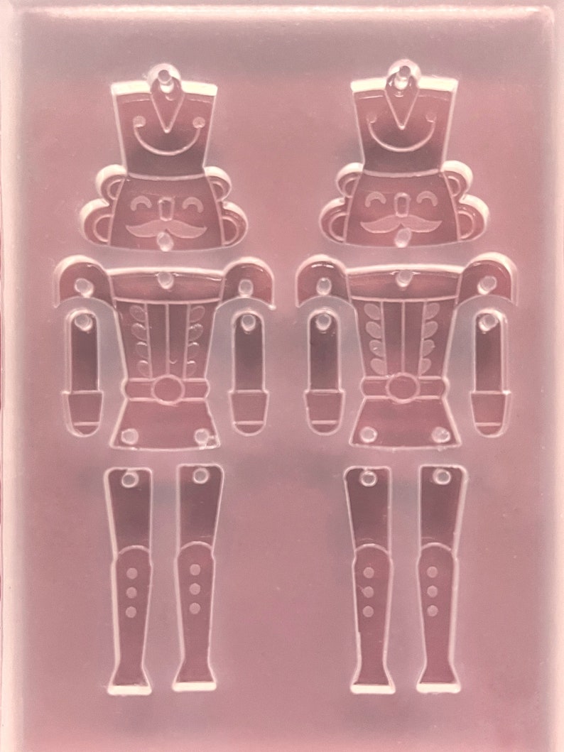 May include: A clear, flexible mould featuring two detailed nutcracker soldier designs. Each soldier has a hat, moustache, and uniform, with separate leg pieces. The mould is designed for crafting decorative items.