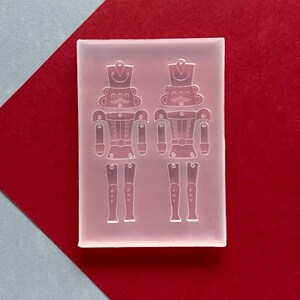 May include: A clear silicone mould featuring two detailed nutcracker soldier designs. The mould is rectangular and sits on a red and grey background. The nutcrackers have hats, moustaches, and detailed uniforms, ideal for crafting projects.