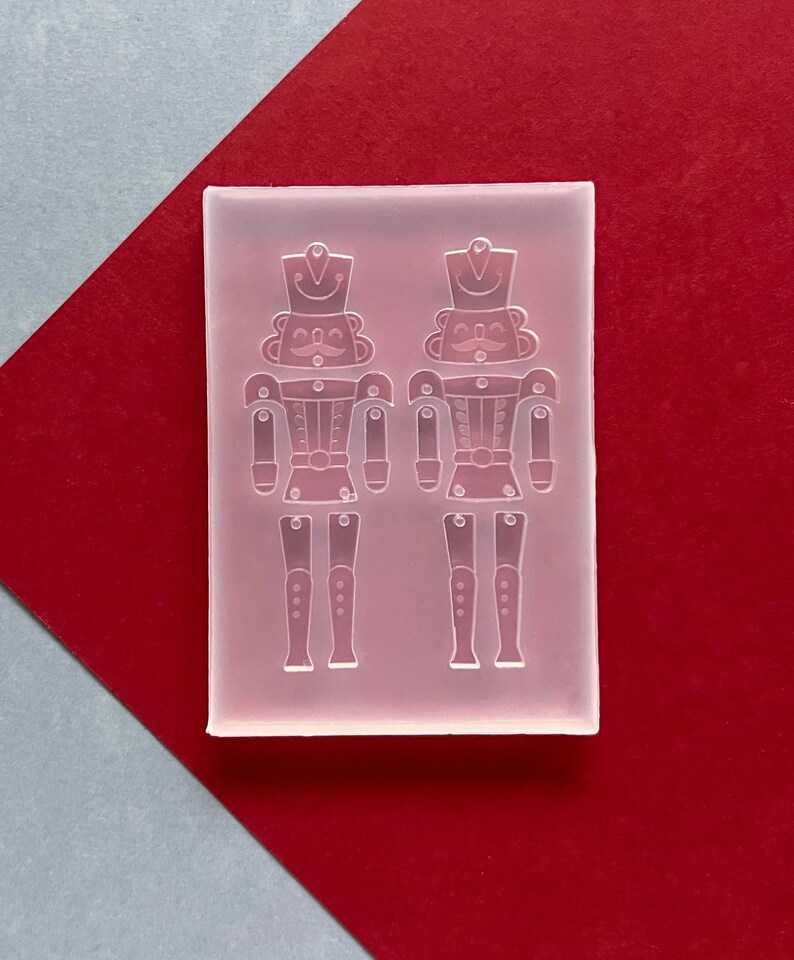 May include: A clear silicone mould featuring two detailed nutcracker soldier designs. The mould is rectangular and sits on a red and grey background. The nutcrackers have hats, moustaches, and detailed uniforms, ideal for crafting projects.
