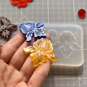Peut inclure: Une main tient deux pièces de résine transparentes en forme de fleur, une bleue et une jaune. D'autres fleurs en résine rouges, transparentes et marron sont dispersées sur une surface grise. Un moule en silicone en forme de fleur est également visible.