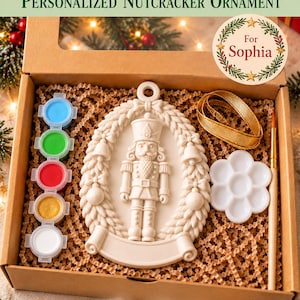 May include: A DIY painting gift kit featuring a white nutcracker ornament inside a cardboard box. The kit includes five small paint pots in blue, green, red, gold, and white, a white flower-shaped paint palette, a paintbrush, and a gold ribbon.