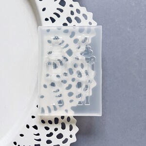 May include: A clear silicone mould in a rectangular shape, featuring two detailed nutcracker soldier designs. The mould is for crafting, with the nutcracker figures visible in relief. The mould is placed on a white, decorative doily.
