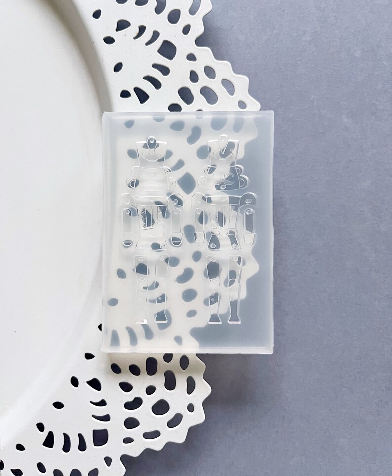 May include: A clear silicone mould in a rectangular shape, featuring two detailed nutcracker soldier designs. The mould is for crafting, with the nutcracker figures visible in relief. The mould is placed on a white, decorative doily.