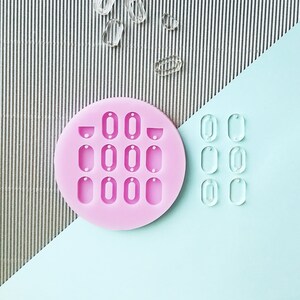 Puede incluir: Un molde de silicona rosa con 12 cavidades ovaladas, cada una con un peque&ntilde;o agujero en la parte superior. Hay 12 &oacute;valos de pl&aacute;stico transparente al lado del molde.