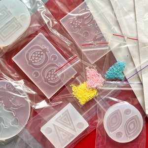 Puede incluir: Surtido de moldes de silicona transparentes para manualidades. Los moldes tienen forma de unicornio, granada, dise&ntilde;o geom&eacute;trico y c&iacute;rculo con un patr&oacute;n de espiral. Los moldes se muestran sobre una superficie roja con cuentas de colores.