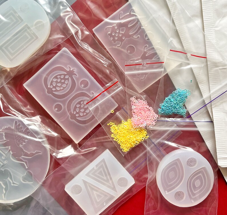 Puede incluir: Surtido de moldes de silicona transparentes para manualidades. Los moldes tienen forma de unicornio, granada, dise&ntilde;o geom&eacute;trico y c&iacute;rculo con un patr&oacute;n de espiral. Los moldes se muestran sobre una superficie roja con cuentas de colores.