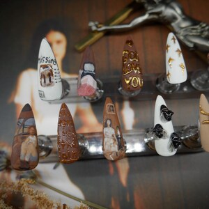May include: A set of ten almond-shaped, hand-painted artificial nails. Designs include text, abstract patterns, and miniature artwork in various colors. One nail reads "Missing". The nails are displayed on clear stands.