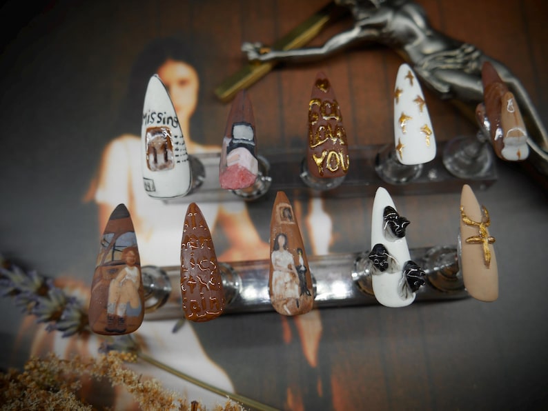 May include: A set of ten almond-shaped, hand-painted artificial nails. Designs include text, abstract patterns, and miniature artwork in various colors. One nail reads "Missing". The nails are displayed on clear stands.