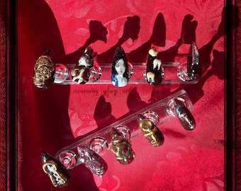 May include: A collection of custom-designed press-on nails displayed on a clear stand. The nails feature various designs, including gold skulls, a cartoon character, a portrait, and abstract patterns. The background is a red fabric with a floral pattern, framed in a dark red ornate frame.