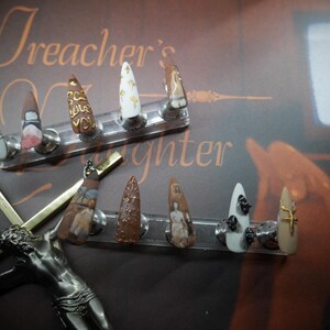 May include: A set of nine custom-designed, pointed acrylic nails. Each nail features unique artwork, including text, patterns, and miniature paintings. The nails are displayed on a clear stand against a brown background with the text "Preacher's Daughter".