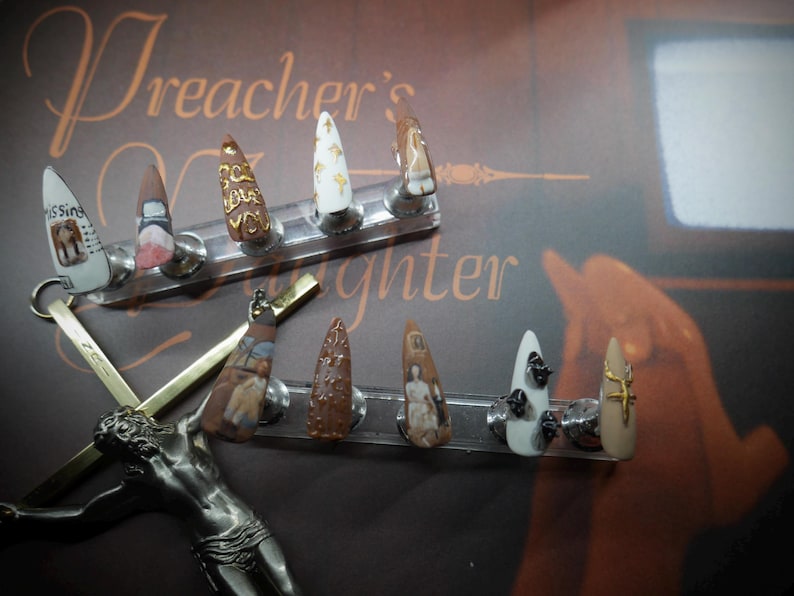 May include: A set of nine custom-designed, pointed acrylic nails. Each nail features unique artwork, including text, patterns, and miniature paintings. The nails are displayed on a clear stand against a brown background with the text "Preacher's Daughter".