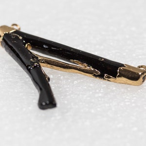 May include: Black and gold pendant with a unique, organic shape. The pendant is made of a black material with gold accents. It has a loop at each end for attaching to a chain or cord.