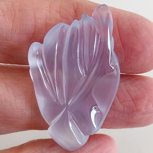 Puede incluir: Una piedra preciosa de calcedonia lavanda tallada en forma de flor. La piedra preciosa es transl&uacute;cida y tiene un acabado liso y pulido.