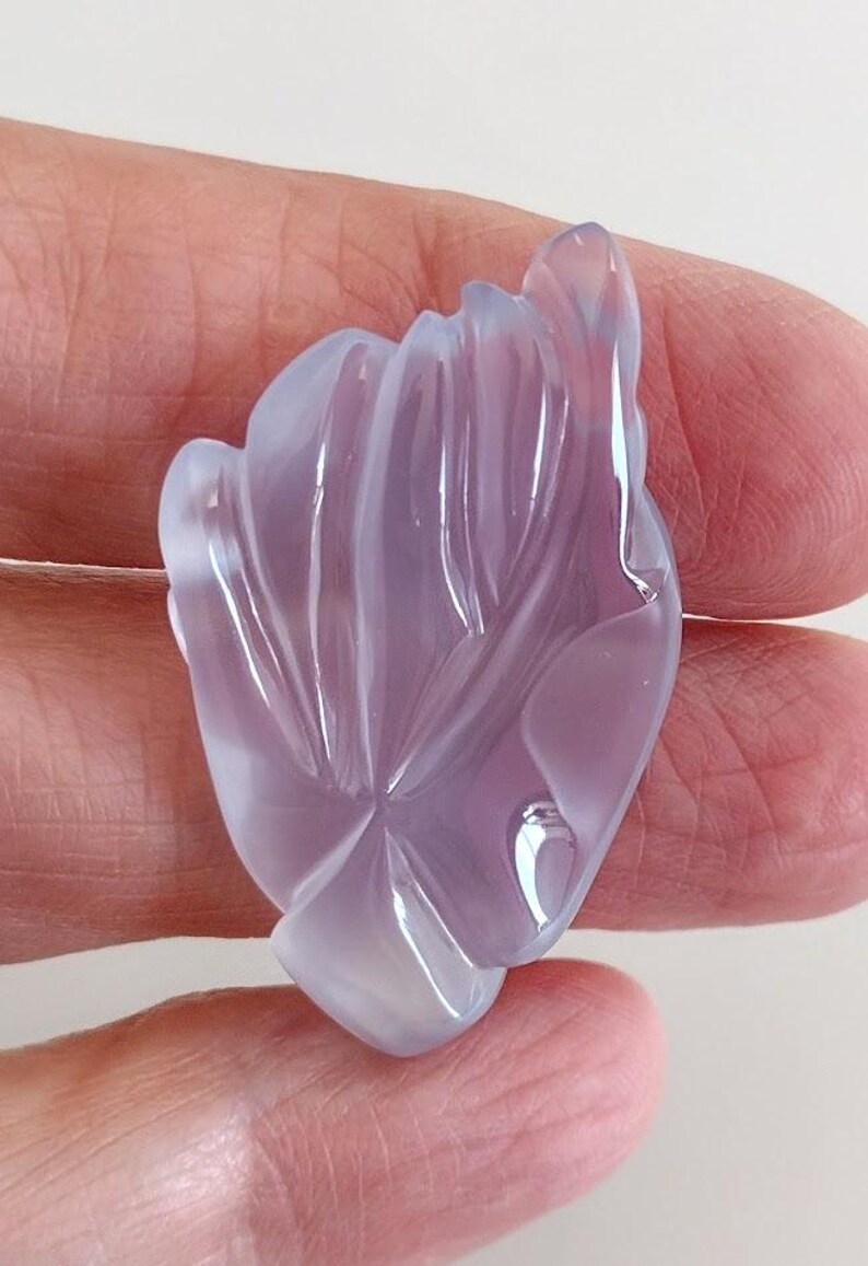 Puede incluir: Una piedra preciosa de calcedonia lavanda tallada en forma de flor. La piedra preciosa es transl&uacute;cida y tiene un acabado liso y pulido.