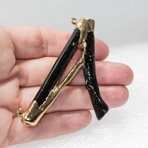 May include: A gold and black letter A pendant charm. The letter is made of a black material with gold accents. The pendant has two loops for attaching to a chain or bracelet.