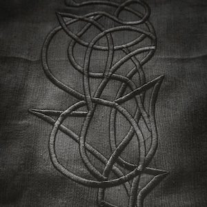 May include: Close-up of a dark gray fabric with an embroidered abstract design. The design features intertwined lines and shapes, creating a textured, monochromatic pattern. The embroidery is in a darker shade of gray.