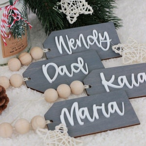 May include: Four rectangular gray wooden gift tags with white 3D lettering reading Wendy, Dad, Kaia, and Maria. Each tag features a string of natural light-colored wooden beads attached to one side, creating a decorative and personalized label set.