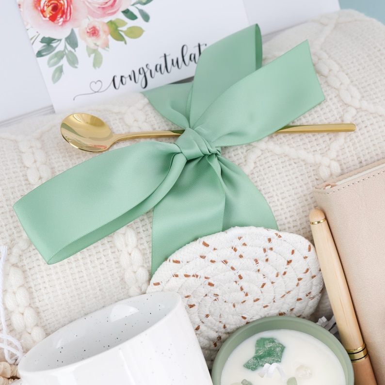 Puede incluir: Un juego de regalo con una tarjeta de felicitaci&oacute;n, una cuchara dorada y un lazo de sat&eacute;n verde claro. El juego incluye una taza blanca, un posavasos tejido, una vela y una cartera beige con un bol&iacute;grafo. La tarjeta tiene detalles florales y la palabra "congratulat".
