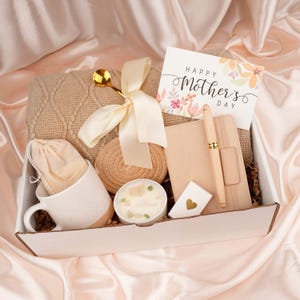 Puede incluir: Una caja de regalo llena de una manta beige de punto trenzado, una taza blanca con base marr&oacute;n, una vela, un cuaderno, un bol&iacute;grafo y una tarjeta que dice "Happy Mother's Day". La caja est&aacute; atada con una cinta color crema.
