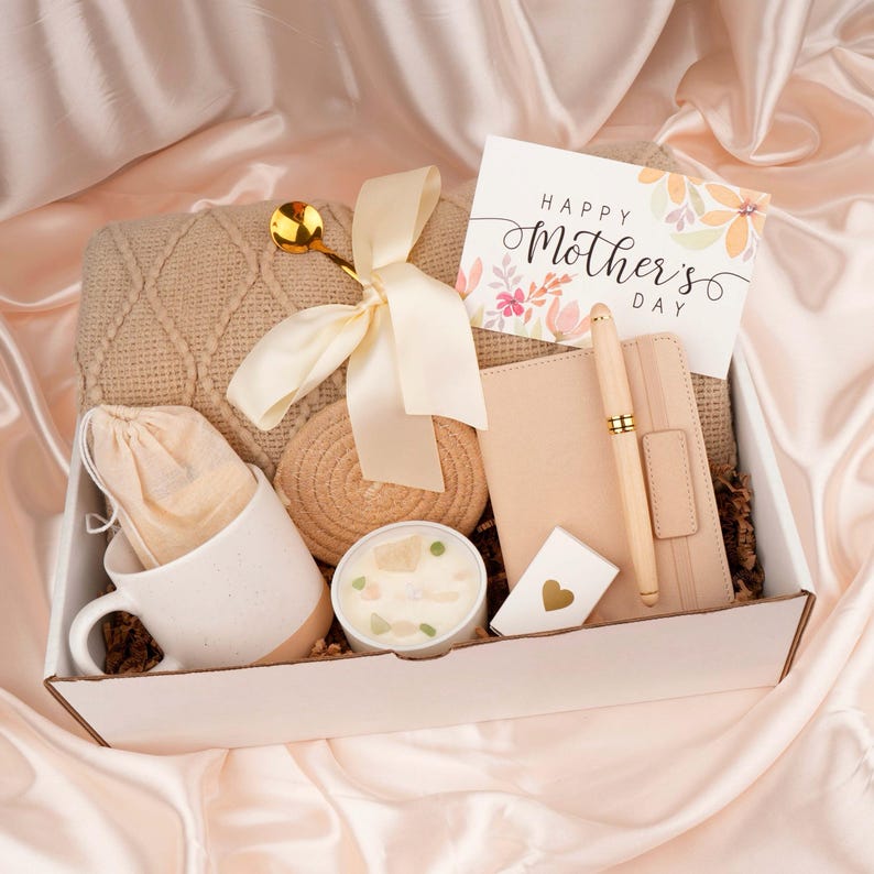 Puede incluir: Una caja de regalo llena de una manta beige de punto trenzado, una taza blanca con base marr&oacute;n, una vela, un cuaderno, un bol&iacute;grafo y una tarjeta que dice "Happy Mother's Day". La caja est&aacute; atada con una cinta color crema.