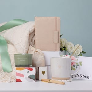 Puede incluir: Un juego de regalo con un diario beige, una taza blanca con un dise&ntilde;o de coraz&oacute;n, una vela perfumada, un bol&iacute;grafo y una caja decorativa. Tambi&eacute;n se incluye una manta blanca y una tarjeta de felicitaci&oacute;n con la palabra "Enhorabuena". Los art&iacute;culos est&aacute;n dispuestos sobre una superficie blanca.