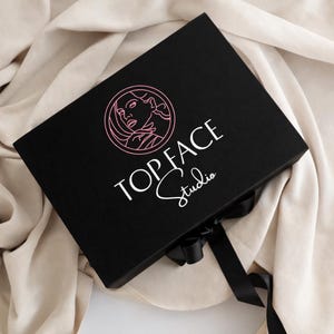 May include: A black box with the text "TOPFACE Studio" in white script and a pink logo of a woman's face. The box is tied with a black ribbon and sits on a beige fabric.