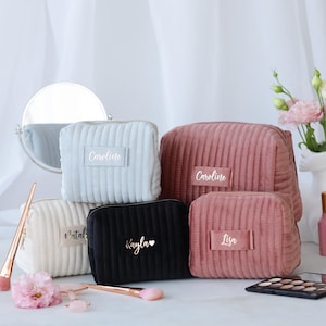 May include: A collection of corduroy cosmetic bags in various colors: white, light blue, black, and pink. Each bag has a rectangular label with a name printed on it. Makeup brushes and a mirror are also visible.