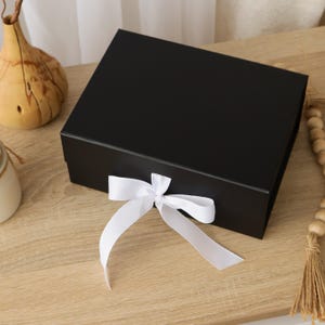 May include: A rectangular black gift box with a smooth matte finish, featuring a white satin ribbon tied in a bow at the front center. The box has a hinged lid design and is presented as a solid, opaque container for storage or presentation purposes.