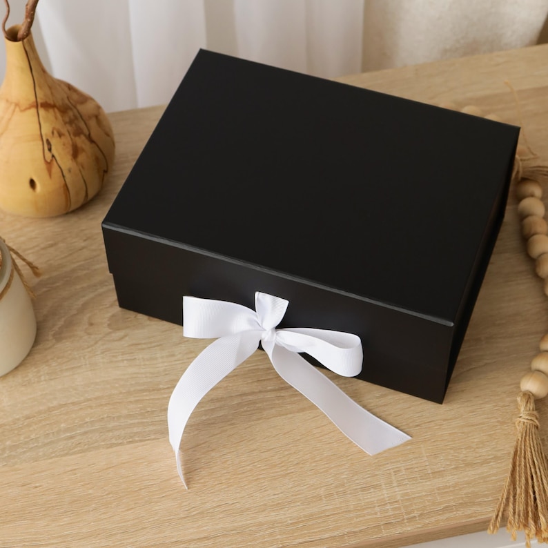 May include: A rectangular black gift box with a smooth matte finish, featuring a white satin ribbon tied in a bow at the front center. The box has a hinged lid design and is presented as a solid, opaque container for storage or presentation purposes.