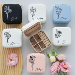 May include: A collection of square jewelry boxes in various colors, including black, white, and light blue. Each box features a floral design and a name in script. One box is open, revealing rings and compartments.