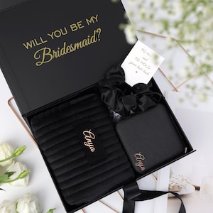 May include: A black gift box with the gold text Will You Be My Bridesmaid? inside the lid. The box contains a black ribbed pouch with the name Anya in gold, a black satin scrunchie, a black square case with Anya in gold, and a white card.