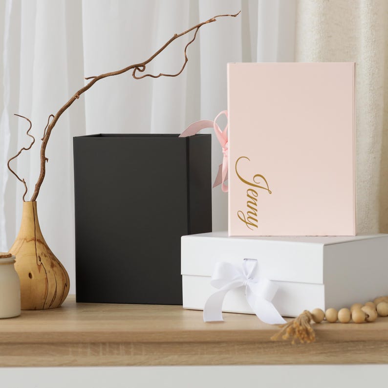 Pode incluir: Tr&ecirc;s caixas de presente: uma preta, uma branca com uma fita branca e uma rosa com o nome "Jenny" em letras douradas. Um galho decorativo e um vaso est&atilde;o no fundo.