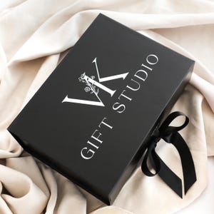 May include: A black gift box with a white logo that reads "VK GIFT STUDIO". The box is tied with a black satin ribbon. The box is resting on a cream-colored fabric.