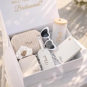 May include: A white gift box with the text Will You Bridesmaid? containing a beige ribbed pouch, white heart-shaped sunglasses, a frosted glass tumbler with a bamboo lid, a white luggage tag, a white passport holder, and a white scrunchie, all personalized with the name Nicole.