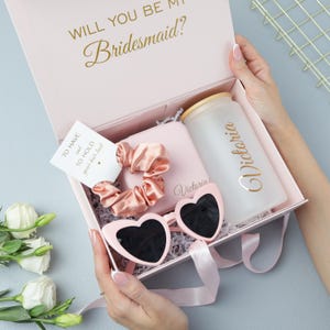 May include: A pink gift box with the text "WILL YOU BE MY Bridesmaid?" contains a scrunchie, heart-shaped sunglasses, a personalized tumbler, and a small card. The box is held open, revealing the contents.