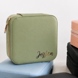May include: A square sage green jewelry case with a textured finish and a gold-toned zipper. The name Jessica is printed in a cursive gold font on the lower right corner of the front panel. The case has rounded corners and a structured design.