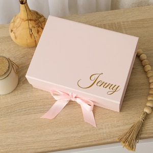 May include: A rectangular light pink gift box featuring a hinged lid and a decorative light pink satin ribbon tied in a bow at the front. The name Jenny is printed on the top right corner of the lid in a metallic gold cursive script font.