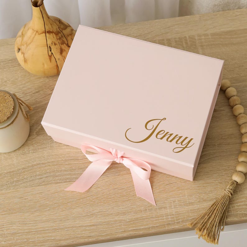 May include: A rectangular light pink gift box featuring a hinged lid and a decorative light pink satin ribbon tied in a bow at the front. The name Jenny is printed on the top right corner of the lid in a metallic gold cursive script font.