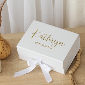 May include: A white gift box with a gold script font that reads "Kathryn" and "Bridesmaid". The box is tied with a white ribbon. The box is on a light wood surface.