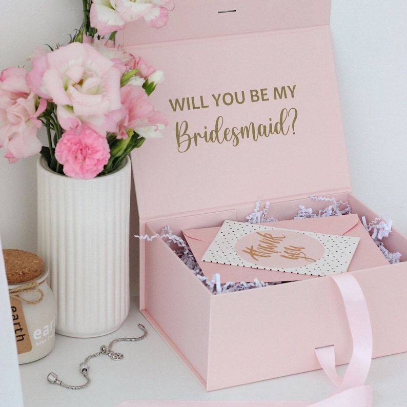 Pode incluir: Caixa de presente rosa claro com texto dourado "WILL YOU BE MY Bridesmaid?". Dentro, um envelope rosa e um cart&atilde;o com "Thank You" s&atilde;o vis&iacute;veis. Um buqu&ecirc; de flores rosas est&aacute; ao lado da caixa. H&aacute; tamb&eacute;m uma pulseira de prata.