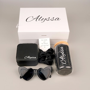 May include: A white gift box with the name "Alyssa" in cursive, alongside a black jewelry case, heart-shaped sunglasses, a jar of hair ties, and a black scrunchie. A small card reads "To have and to hold your hair back."