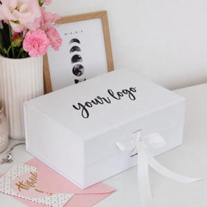 May include: A rectangular white gift box with a magnetic closure and a white ribbon tie. The lid features the black text "your logo" in a cursive script. The box is positioned on a surface with a pink envelope and a framed moon phase print.