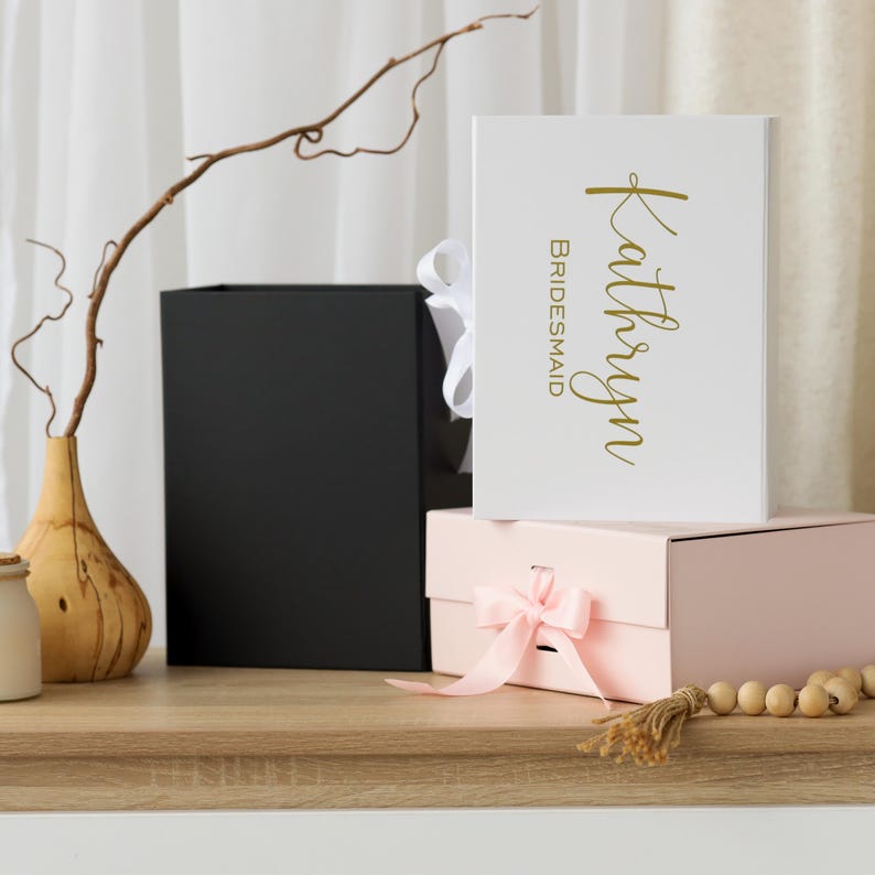 Pode incluir: Uma caixa de presente branca com a inscri&ccedil;&atilde;o dourada "Kathryn Bridesmaid" est&aacute; em cima de uma caixa rosa amarrada com uma fita rosa. Uma caixa preta e um vaso de madeira com um galho tamb&eacute;m s&atilde;o vis&iacute;veis.