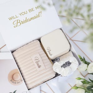 May include: A white gift box with the gold text WILL YOU BE MY BRIDESMAID? containing a beige quilted pouch, a square beige jewelry case, and a white scrunchie. Both the pouch and jewelry case are personalized with the name Nicole in black.