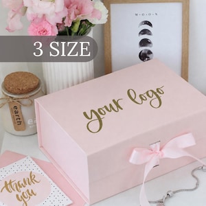 May include: A rectangular light pink gift box with a hinged lid and a matching pink ribbon closure. The top of the box features the text "your logo" in gold script. The box is presented alongside a white card with "thank you" in gold script.