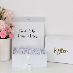 Pode incluir: Caixa de presente branca com o texto "Bride to be! Miss to Mrs." na tampa interna. A caixa est&aacute; aberta e preenchida com peda&ccedil;os de papel branco. Uma segunda caixa branca com o nome "Kylee" em dourado est&aacute; ao lado.