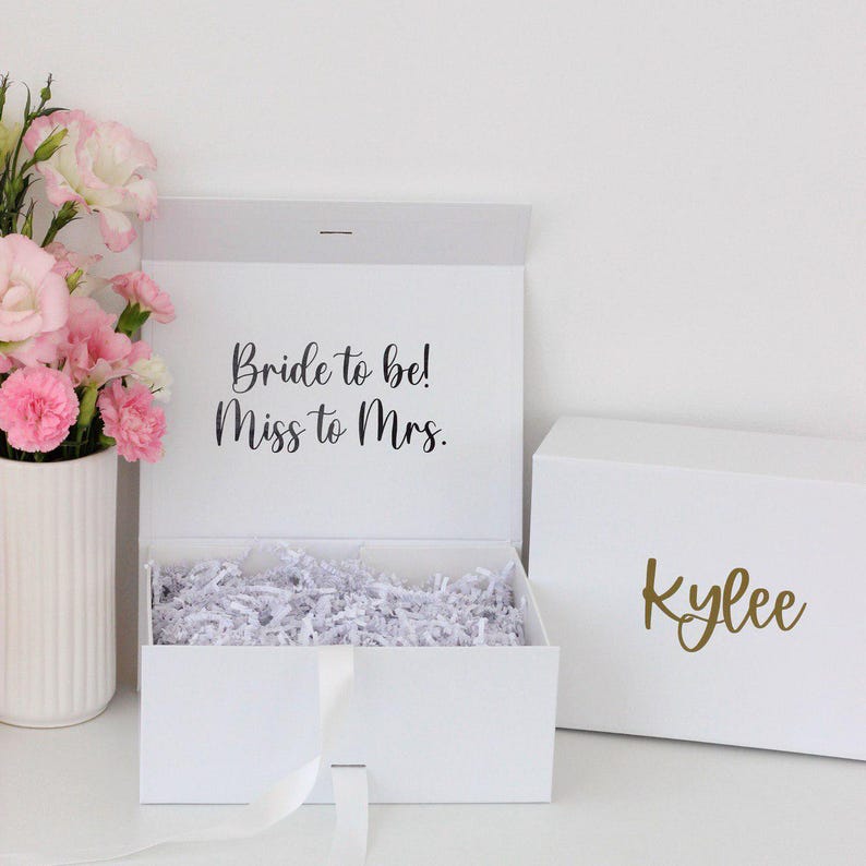 Pode incluir: Caixa de presente branca com o texto "Bride to be! Miss to Mrs." na tampa interna. A caixa est&aacute; aberta e preenchida com peda&ccedil;os de papel branco. Uma segunda caixa branca com o nome "Kylee" em dourado est&aacute; ao lado.