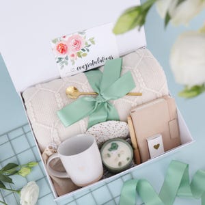 Puede incluir: Una caja de regalo blanca llena de una manta color crema, una taza, una vela, un cuaderno y una cuchara dorada. Tambi&eacute;n se incluye una tarjeta con la palabra "felicidades" y una cinta verde. La taza y la tarjeta tienen un dise&ntilde;o de coraz&oacute;n.