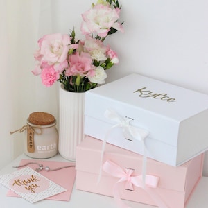 May include: A still-life arrangement featuring a white gift box with the name "Kylee" in gold script, stacked atop a pink box. A vase of pink and white flowers sits nearby, with a small jar and a "thank you" card.