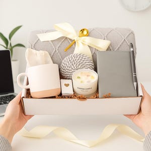 Puede incluir: Una caja de regalo llena de una manta gris, una taza blanca y beige, una vela, un cuaderno, un bol&iacute;grafo, una bola decorativa y una cajita con un coraz&oacute;n. La caja est&aacute; atada con una cinta color crema.
